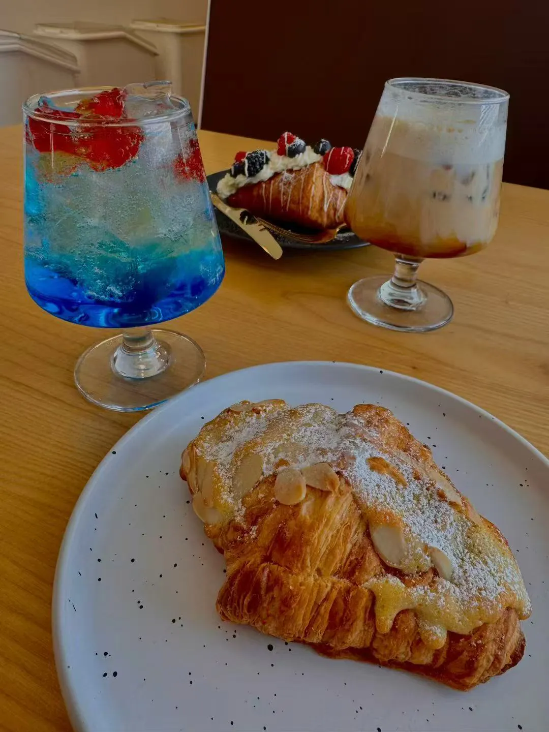 A plated almond croissant with powdered sugar, accompanied by two colorful drinks—one blue layered soda, one iced coffee—and a fruit-topped pastry in the background.