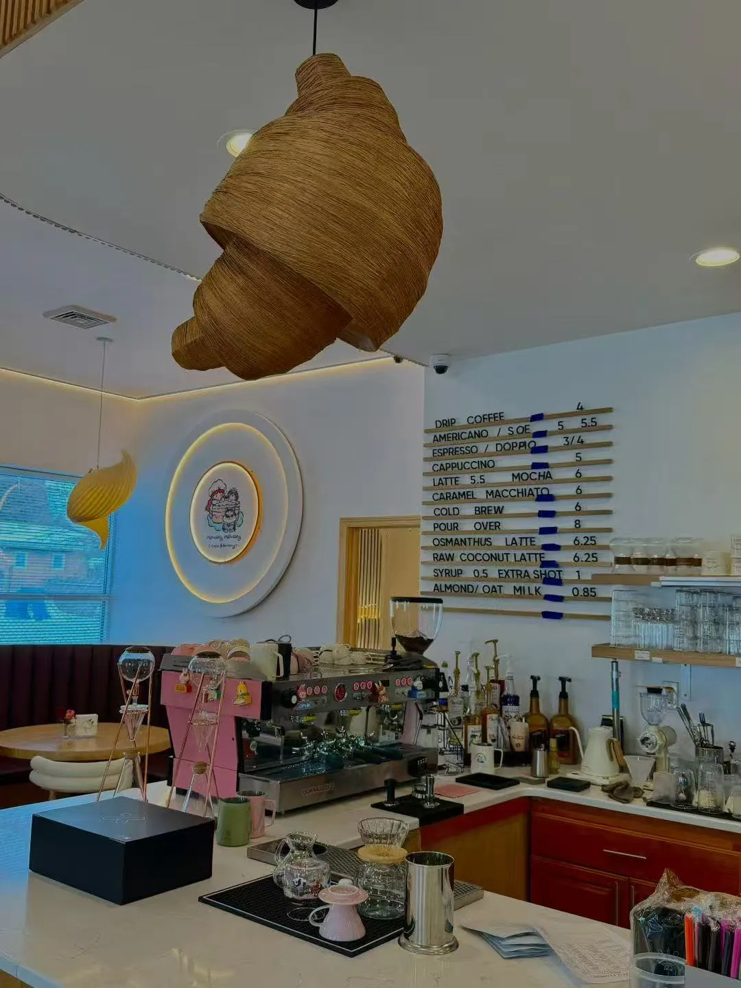 The coffee bar of a stylish café with a giant croissant-shaped pendant light, a menu board, and a pink espresso machine.