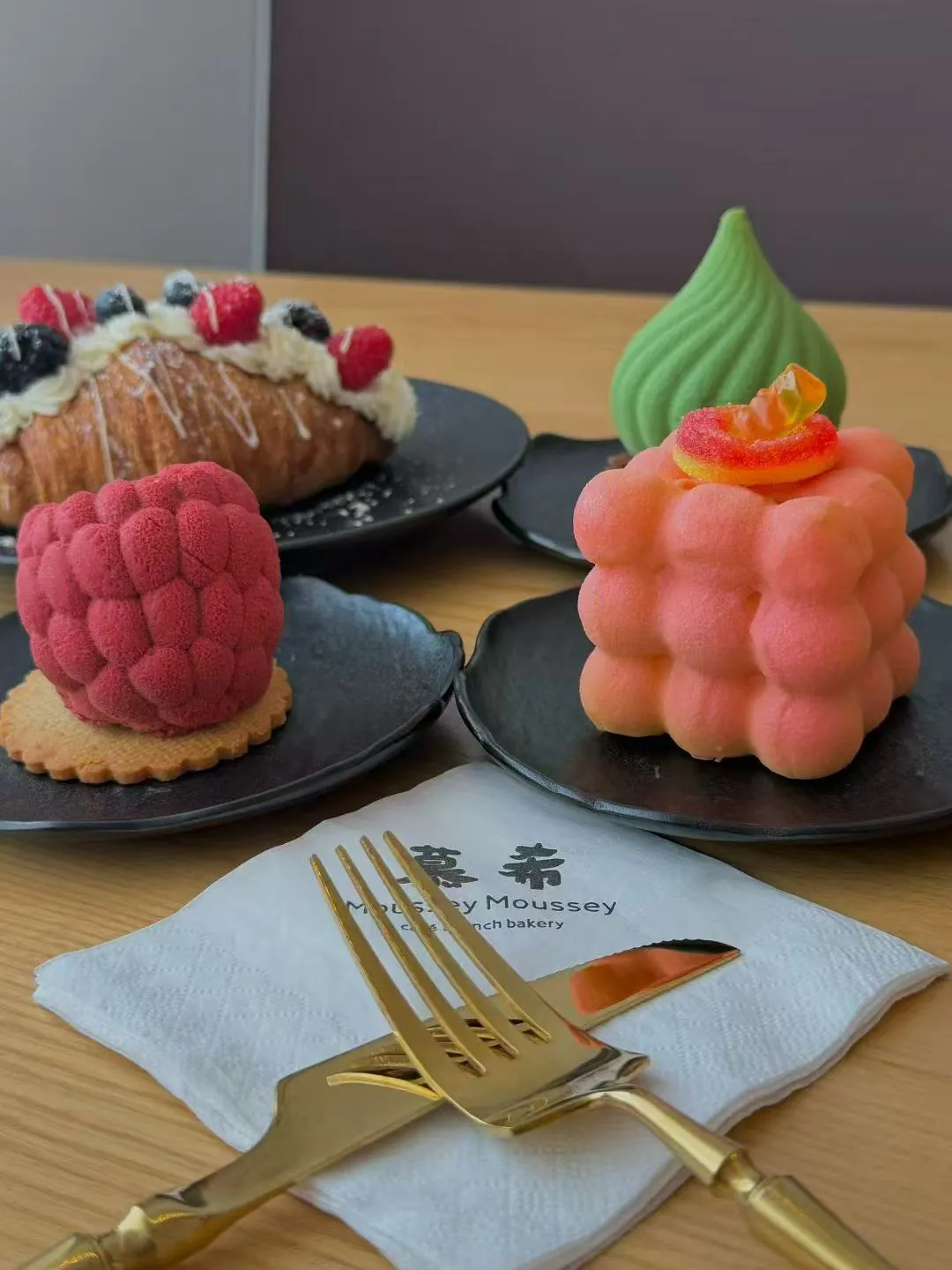 Artistic desserts from Moussy Moussey bakery: a raspberry mousse, a pink bubble-textured cake, and a green dome dessert, served with gold cutlery.