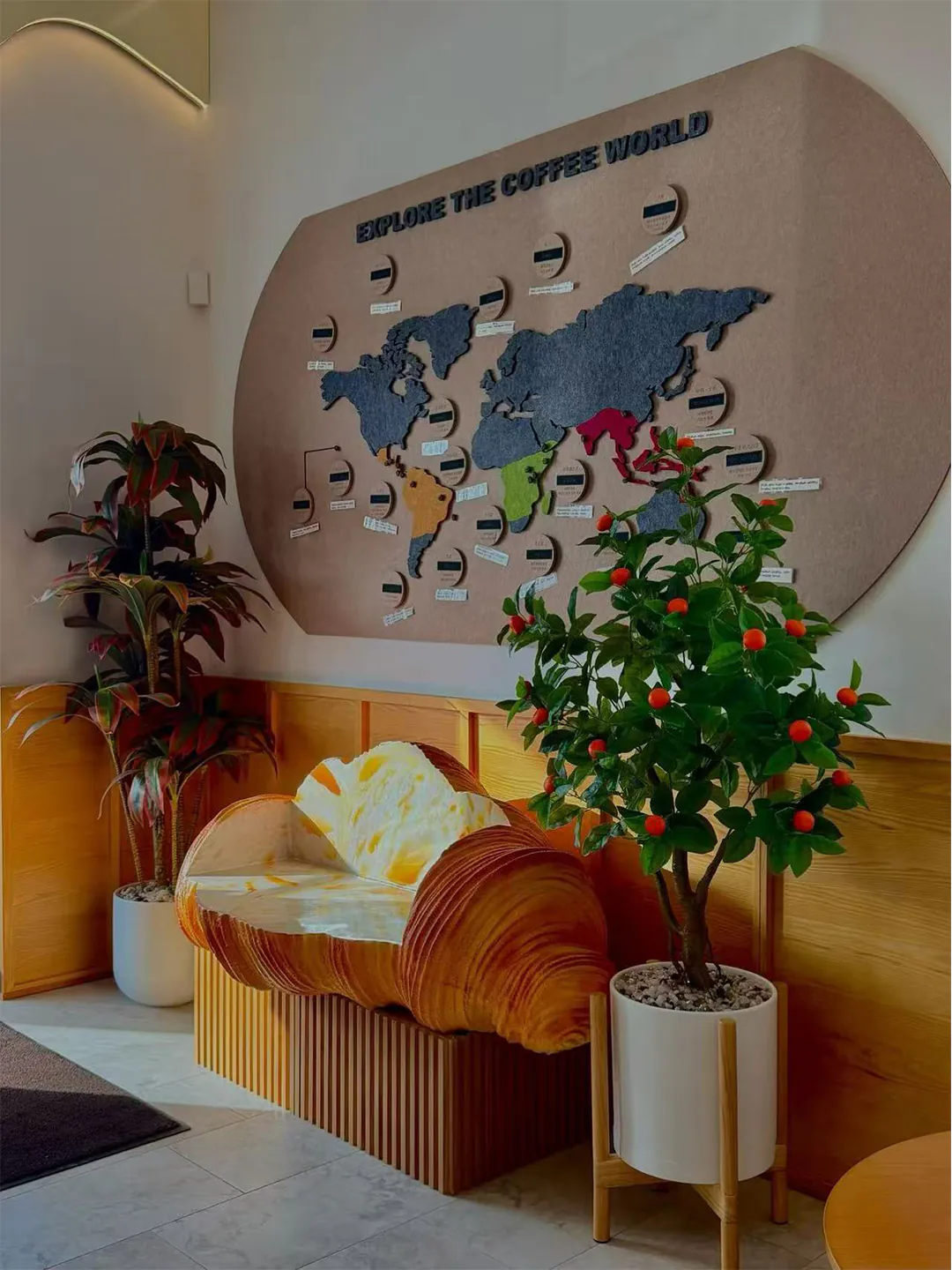 A café corner featuring a large "Explore the Coffee World" world map display, a croissant-shaped bench, and potted plants.