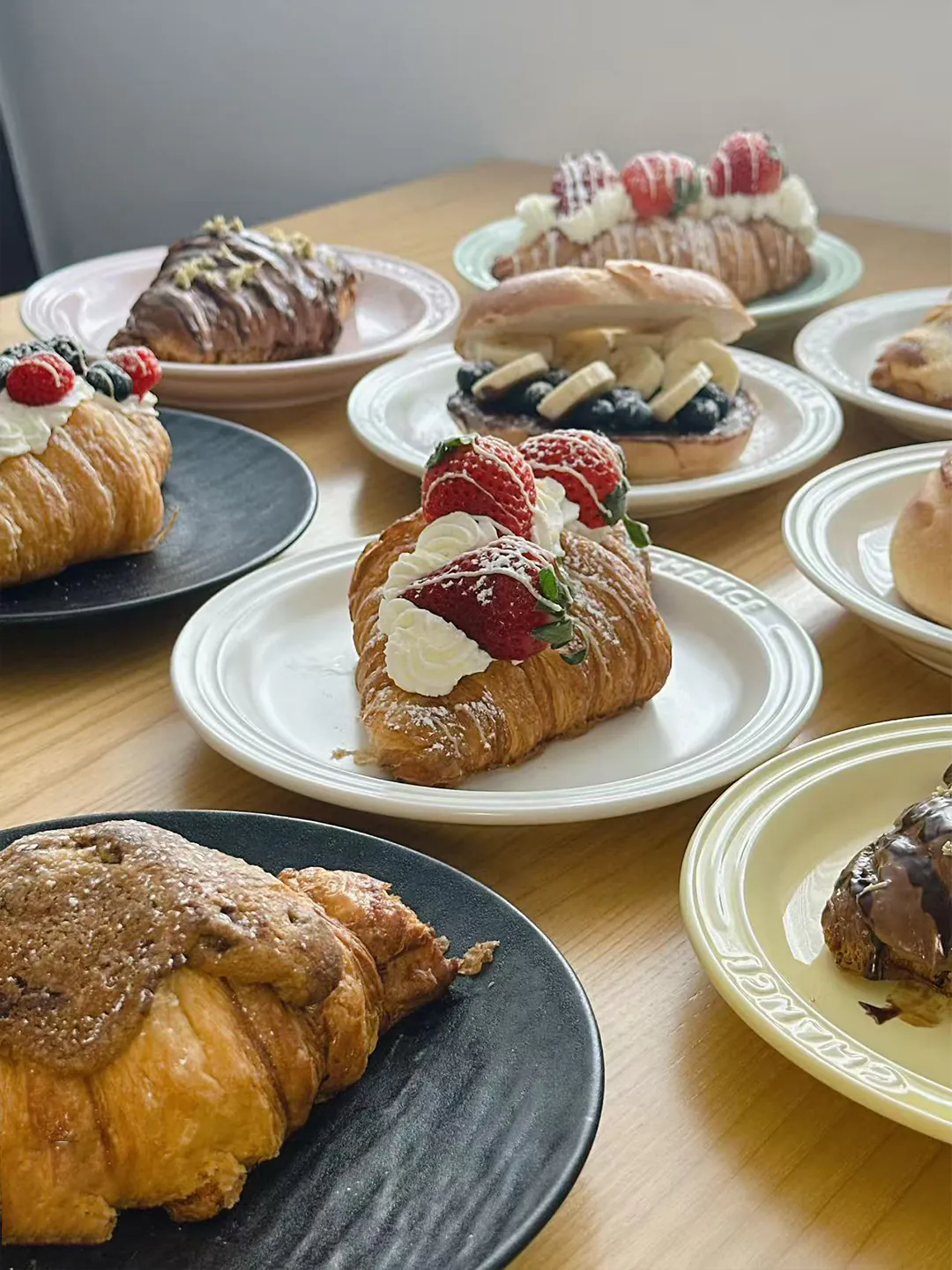 A wooden table laden with assorted pastries including strawberry-topped croissants, chocolate-glazed danishes, and fruit-filled buns on ceramic plates.